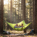 A bright lime green camping hammock suspended between two trees in a sun-dappled pine forest, with three people relaxing inside. Backpacks and a blanket are visible on the forest floor nearby.